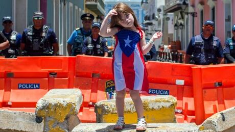 A girl wearing a dress featuring the Puerto Rican flag stands by police blocking the road leading to La Fortaleza, the governor's official residence, in San Juan, Puerto Rico, Thursday, July 18, 2019. Protesters are demanding Gov. Ricardo Rossello resign after the leak of online chats that show him making misogynistic slurs and mocking his constituents. Puerto Rico protests