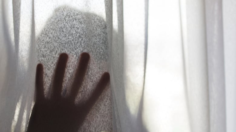 The outline of a person at a window, with their hand on the curtain, seen from the outside of the window.