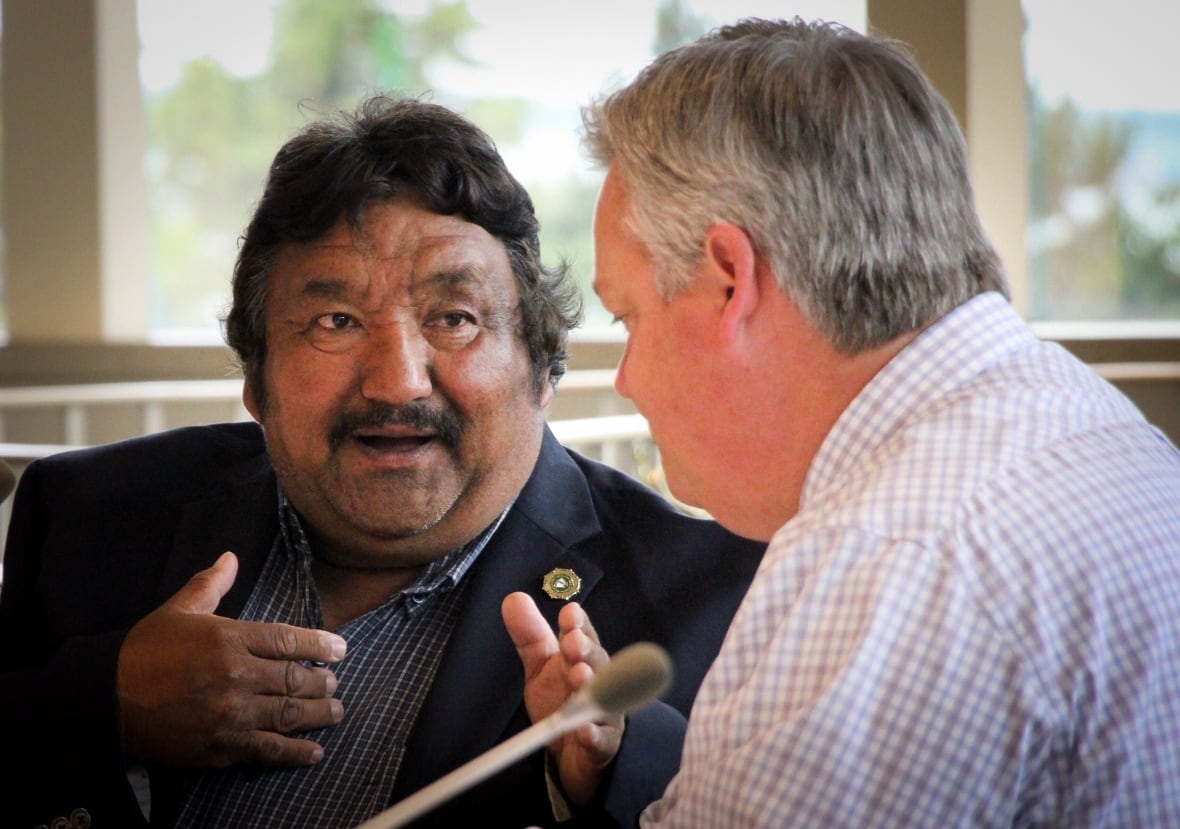 A man with black hair and a dark blue suit gestures with his hands while talking to a man with grey hair and a white checked shirt.