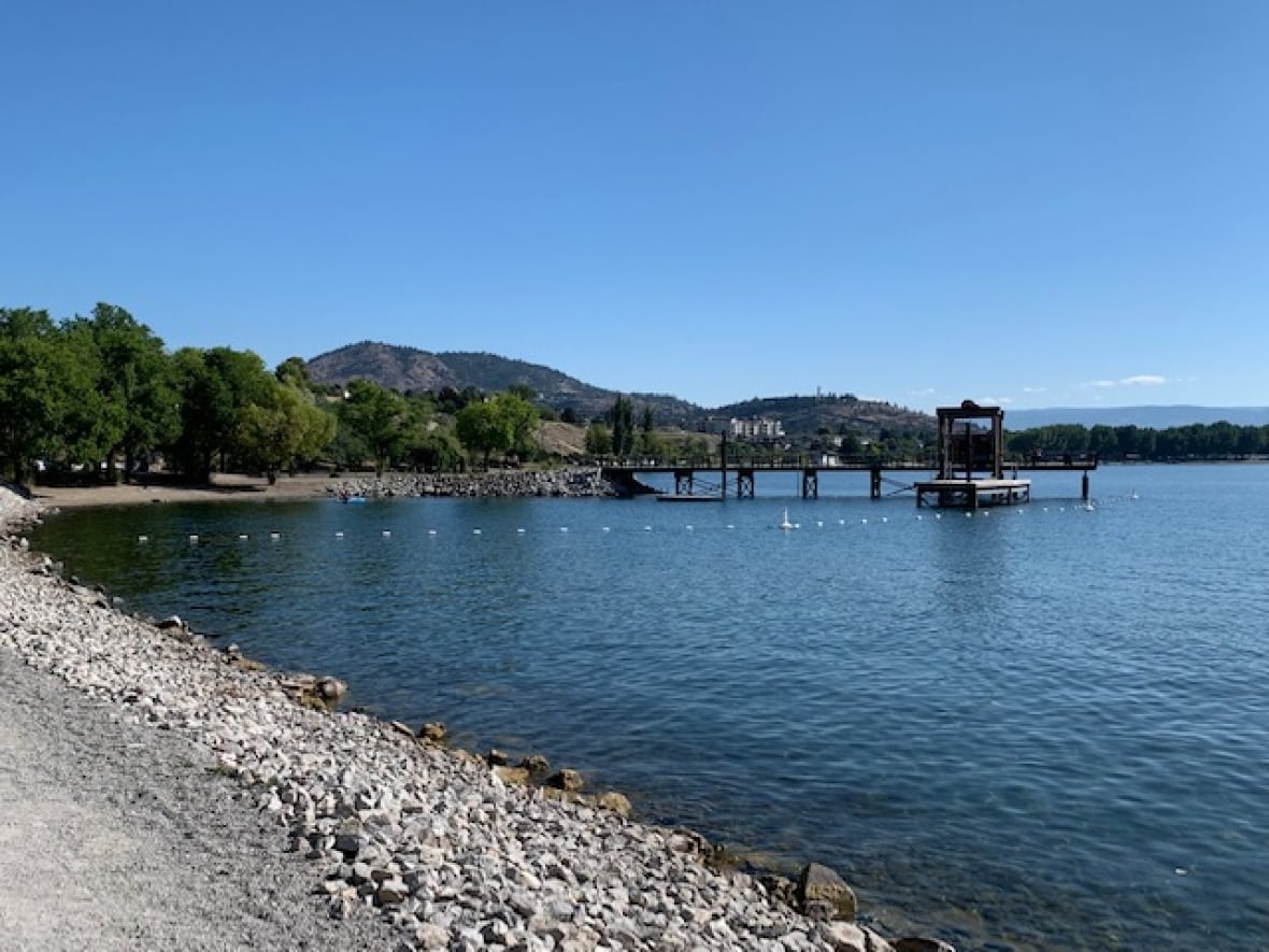 A blue lake with a pier extending into it with riprock in the foreground and green hilly terrain in the background.