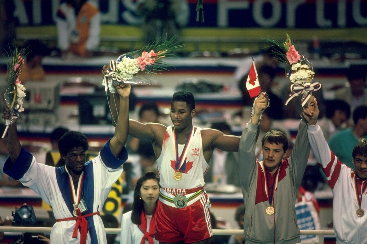 Four men's boxers stand on an Olympic podium in a boxing ring, holding their arms in the air while holding onto bouquets of flowers.