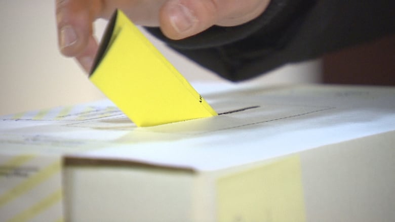 A hand drops a yellow ballot into a white ballot box.