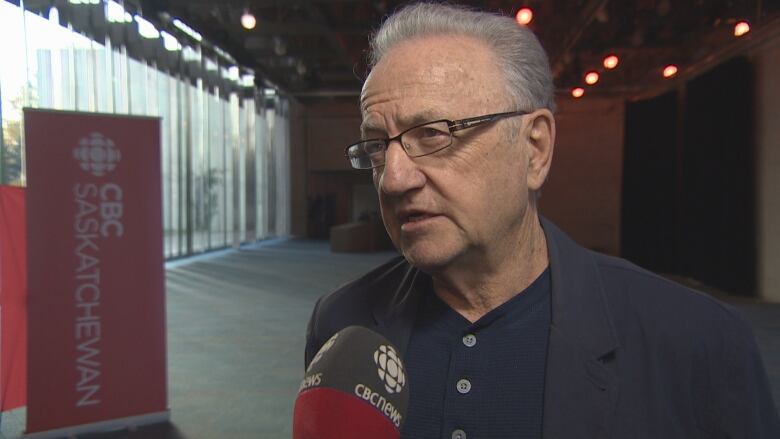 A man with grey hair and a blue shirt and black jacket talks into a microphone. 