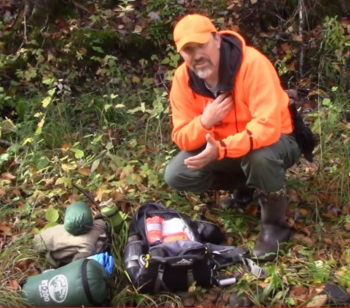 A man in an orange hat and shirt kneeling in the woods with camping gear.