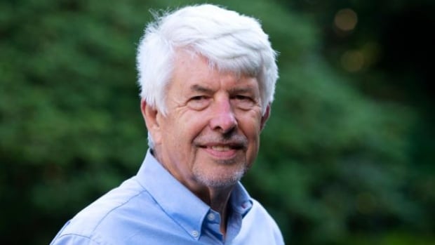 A man with a shock of white hair in a blue open-collared shirt standing in front of blurred-out greenery smiles into the camera,