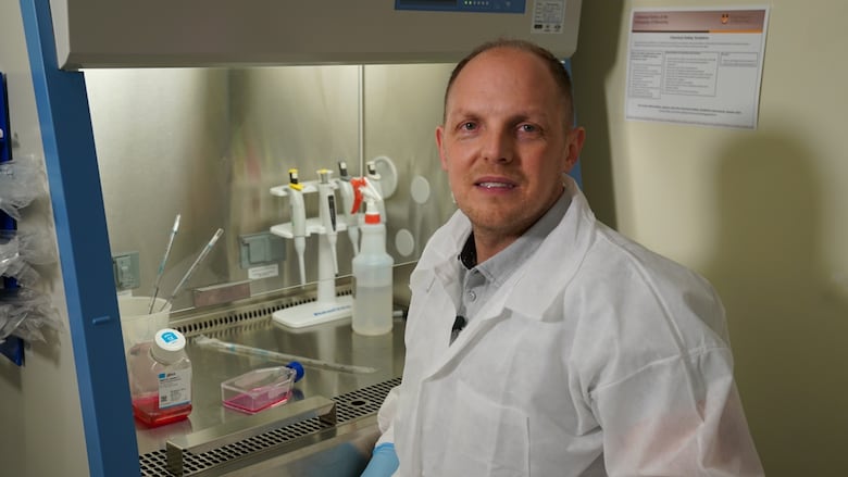 A man in a white lab coat stands in front of lab equipment. 