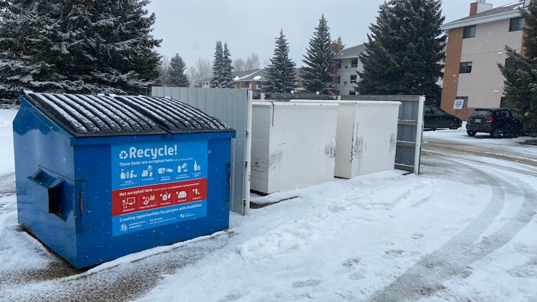 Large blue commercial recycling bin sits near snow-covered road. Spruce trees and an apartment building are in the background