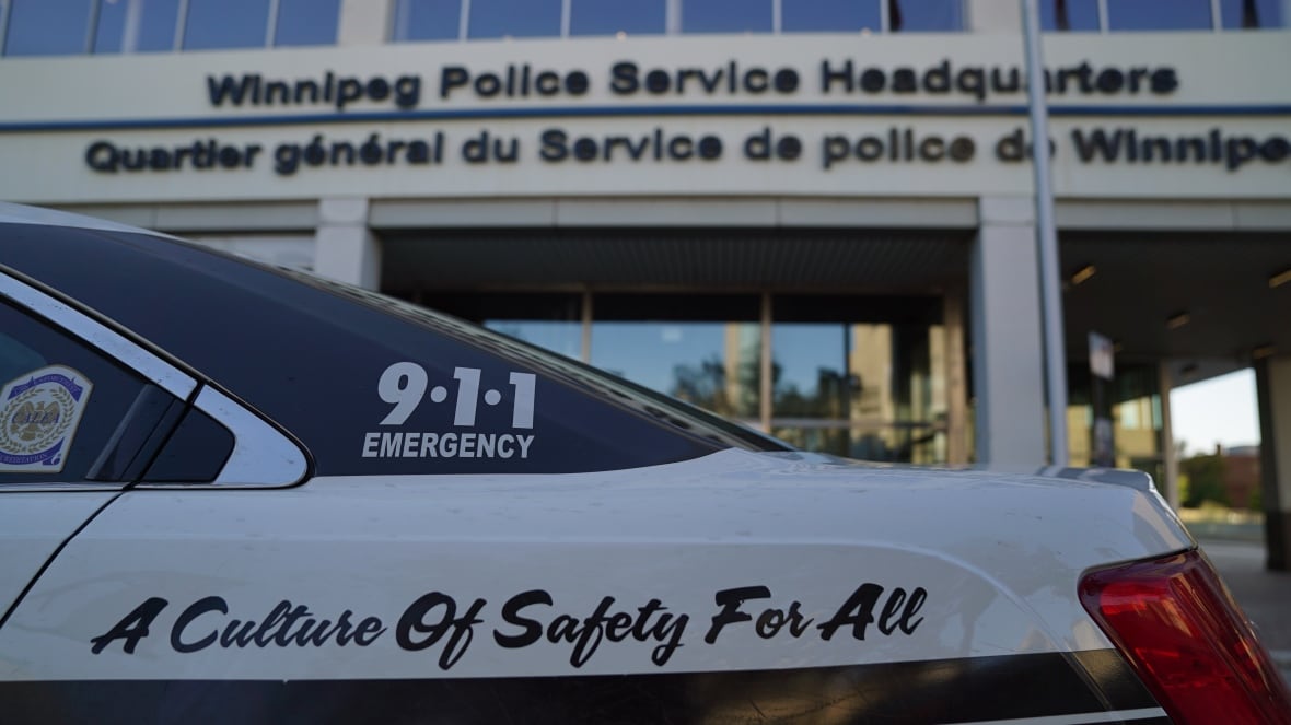 A police cruiser is seen close up in the foreground, with the 911 emergency number on it. The police headquarters building is in the background.