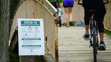 Several people pass a COVID-19 sign beside a bridge in Victoria Park, Kitchener. Bike passing COVID-19 sign Victoria Park Kitchener