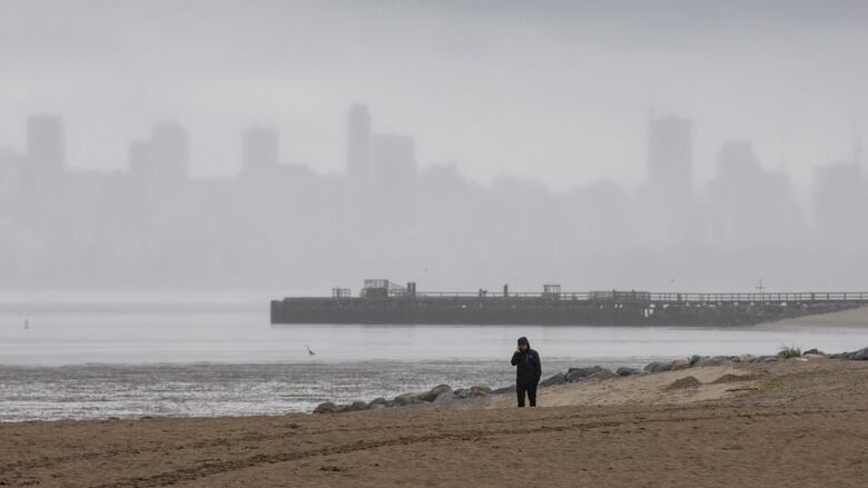 A person wearing a rain jacket walks on a cloudy, rainy beach in Vancouver with the city skyline obscured by fog in the background.