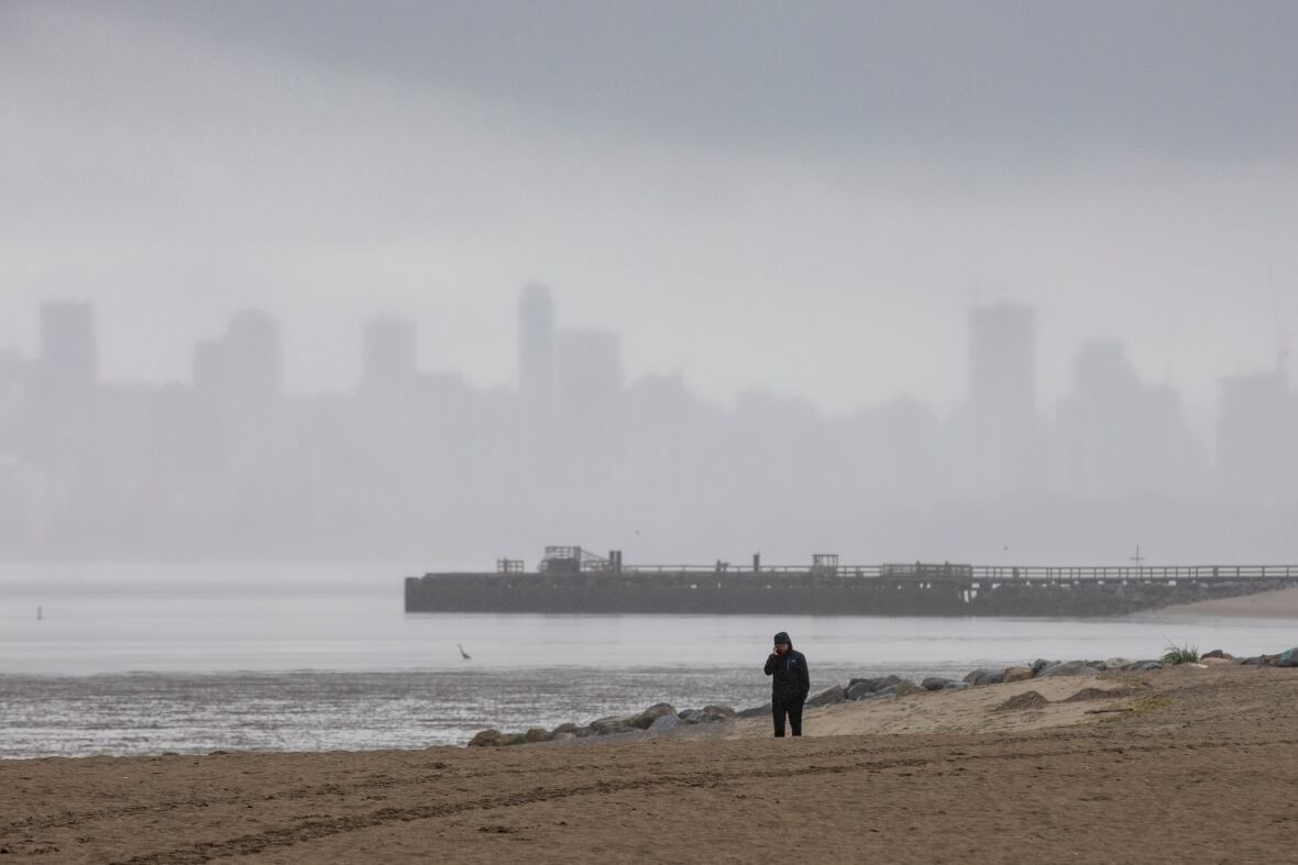 A person wearing a rain jacket walks on a cloudy, rainy beach in Vancouver with the city skyline obscured by fog in the background.