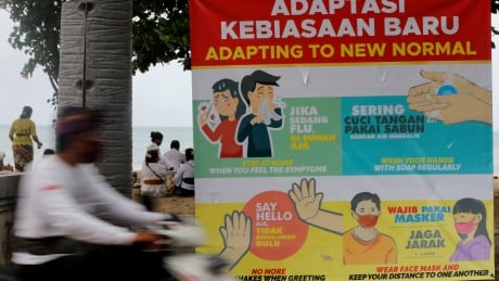 A man rides his motorcycle pass a banner of adapting to coronavirus protection measures at Kuta beach, Bali, Indonesia, Thursday, July 9, 2020. Indonesia's resort island of Bali reopened after a three-month virus lockdown Thursday, allowing local people and stranded foreign tourists to resume public activities before foreign arrivals resume in September. Virus Outbreak Indonesia