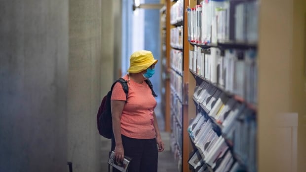 A woman in a pink T-shirt and yellow hat looks at shelves of books at a public library.