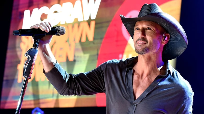 A man with a goatee and black cowboy hat holds a mic while standing on stage.
