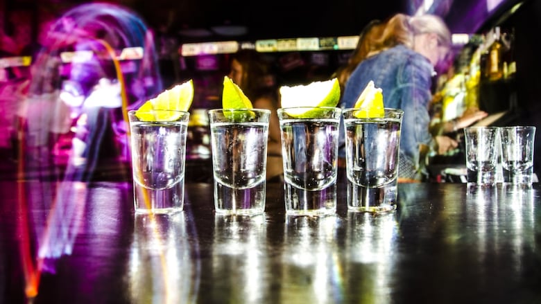 A close-up photograph of four shot glasses lined up on a bar, full of clear liquid with wedges of lemon or lime in them.