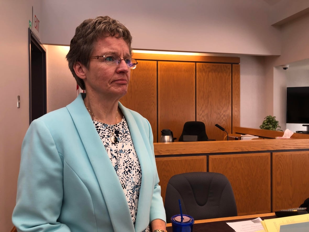 A woman with short hair and glasses, wearing a light blue blazer, stands in front of a wooden judge's bench.