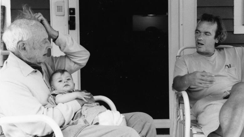 A black and white photo of two men in conversation, one of the men is holding a baby.