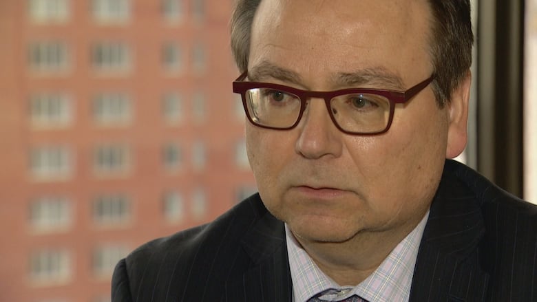 A man with maroon glasses, grey hair and a dark suit is seen up close sitting at a table in an office with a window in the background.