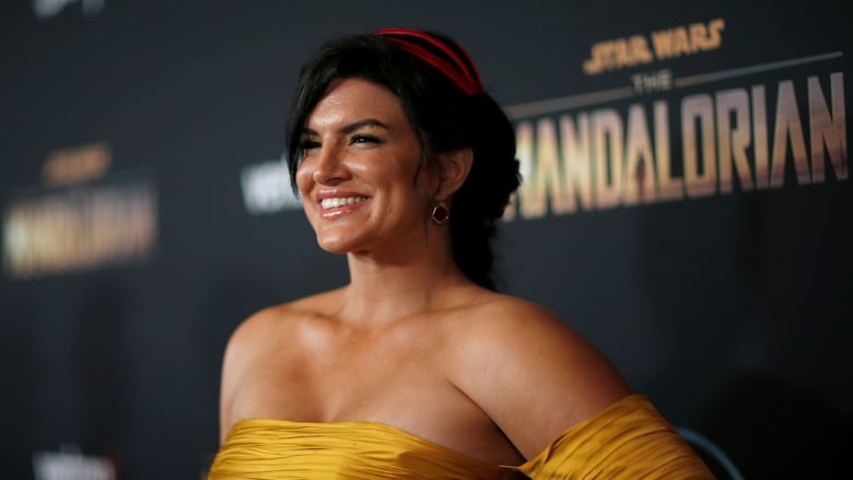 A woman with dark hair and a yellow dress smiles for the camera at a red carpet event. behind her, black posters promoting the show The Mandalorian are visible