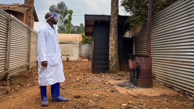 A medical worker wearing personal protective equipment looks at a metal barrel, placed in front of a tree, in front of a steel barrier. 