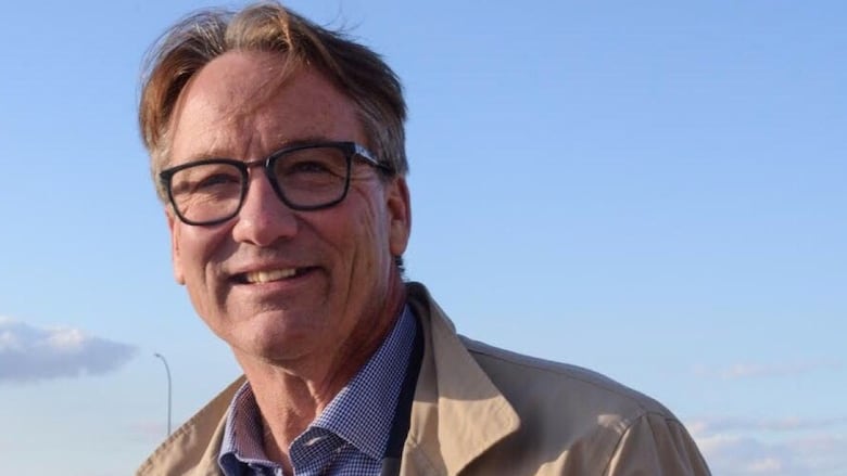 A middle-aged man with dark blond hair and wearing glasses smiles for the camera as he stands outside with a field in the background and a blue sky.