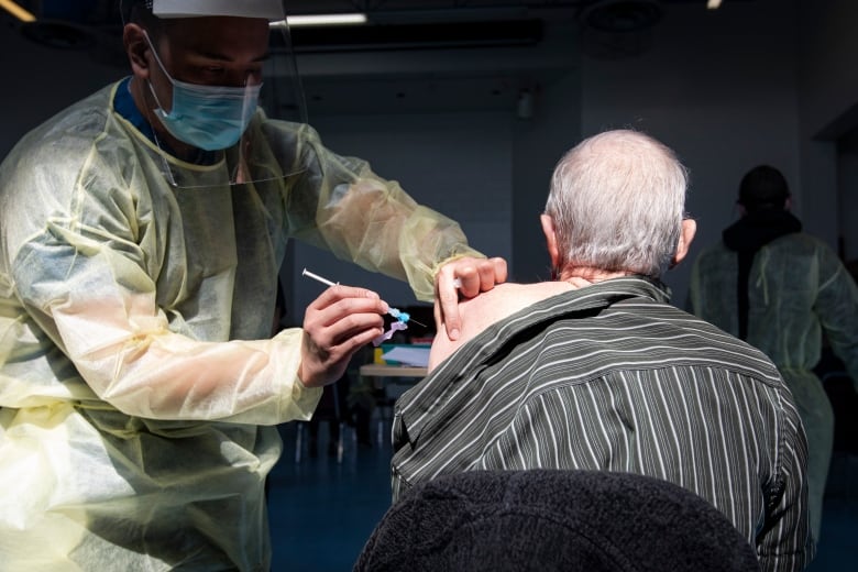 A health-care worker wearing individual protective equipment, including a look shield and mask, administers a vaccine into nan limb of an aged man.