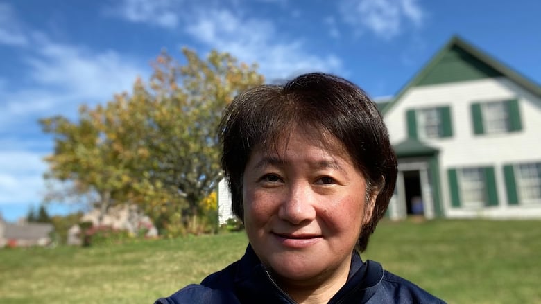 A woman wearing a blue zip up jacket stands in front of a green and white house, a tree is to the left of the image.