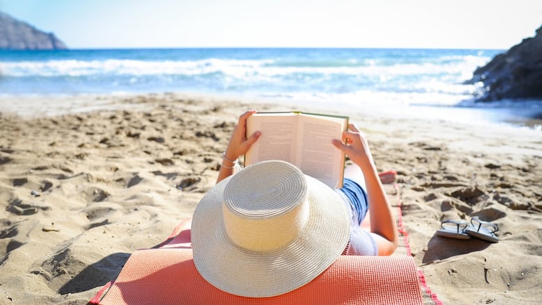 Caucasian woman is reading a book on a sandy beach wearing a straw hat