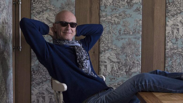 A person in sunglasses leans back on a wooden chair and buts their legs up on a table in front of them.
