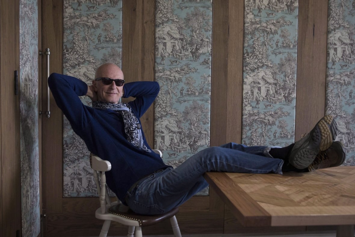 A person in sunglasses leans back on a wooden chair and buts their legs up on a table in front of them.
