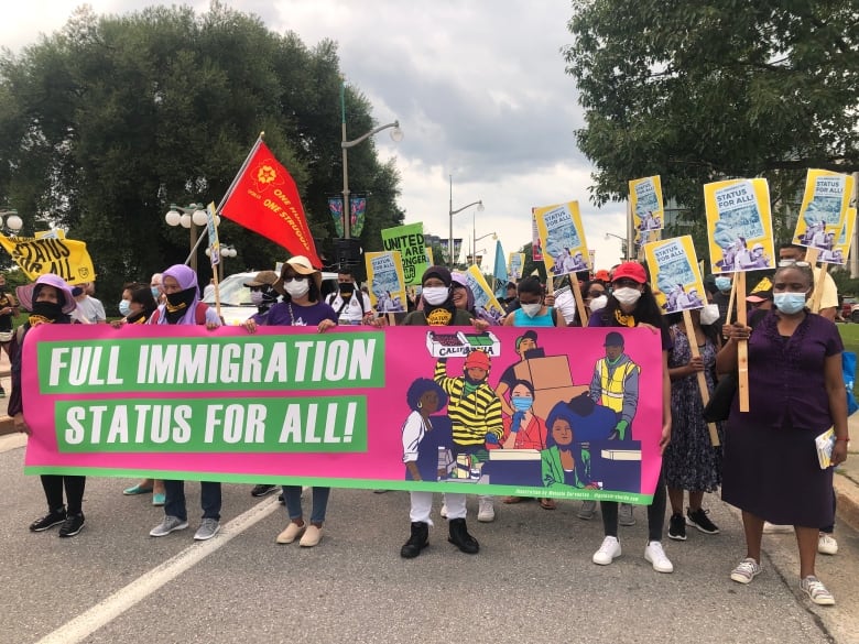 A group of people standing in a street hold placards and march behind a sign reading "'Full immigration status for all!"