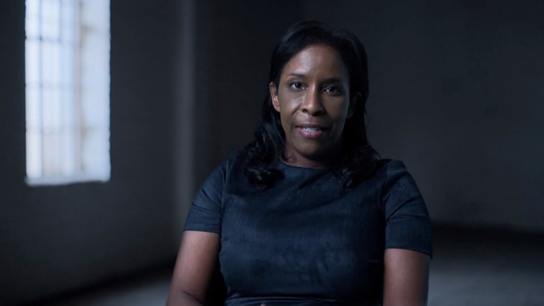 A middle-aged woman sitting in a dark room, talking to camera.