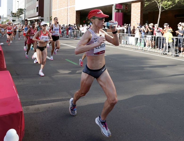 Canadian track and field athlete Malindi Elmore runs in Tokyo during the Olypmic games wearing a red Canada hat and stripped uniform top. She drinks from a water bottle at an air station along the course.