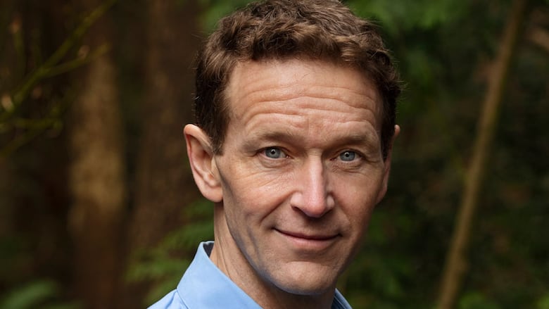 A man with short brown hair poses for a headshot outside, dressed in a light blue dress shirt. 