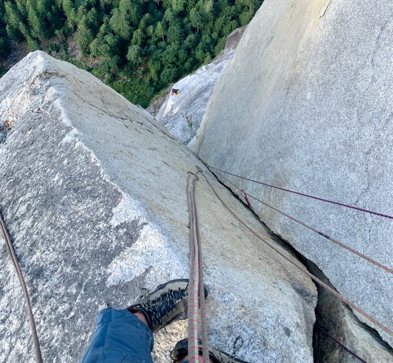 Vertiginous view down a rock slab with trees in the background and climber's feet, ropes in the foreground.