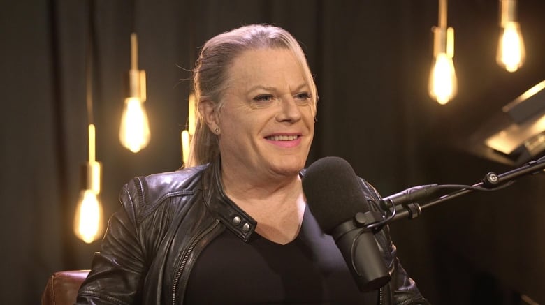 A woman with long blonde hair and a black leather jacket smiles in front of a microphone in a room with decorative hanging lights. 