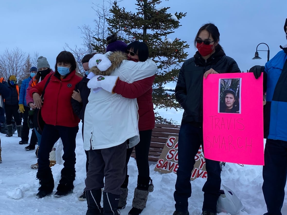 Women standing in the snow holding a picture of a young man, receiving hugs.