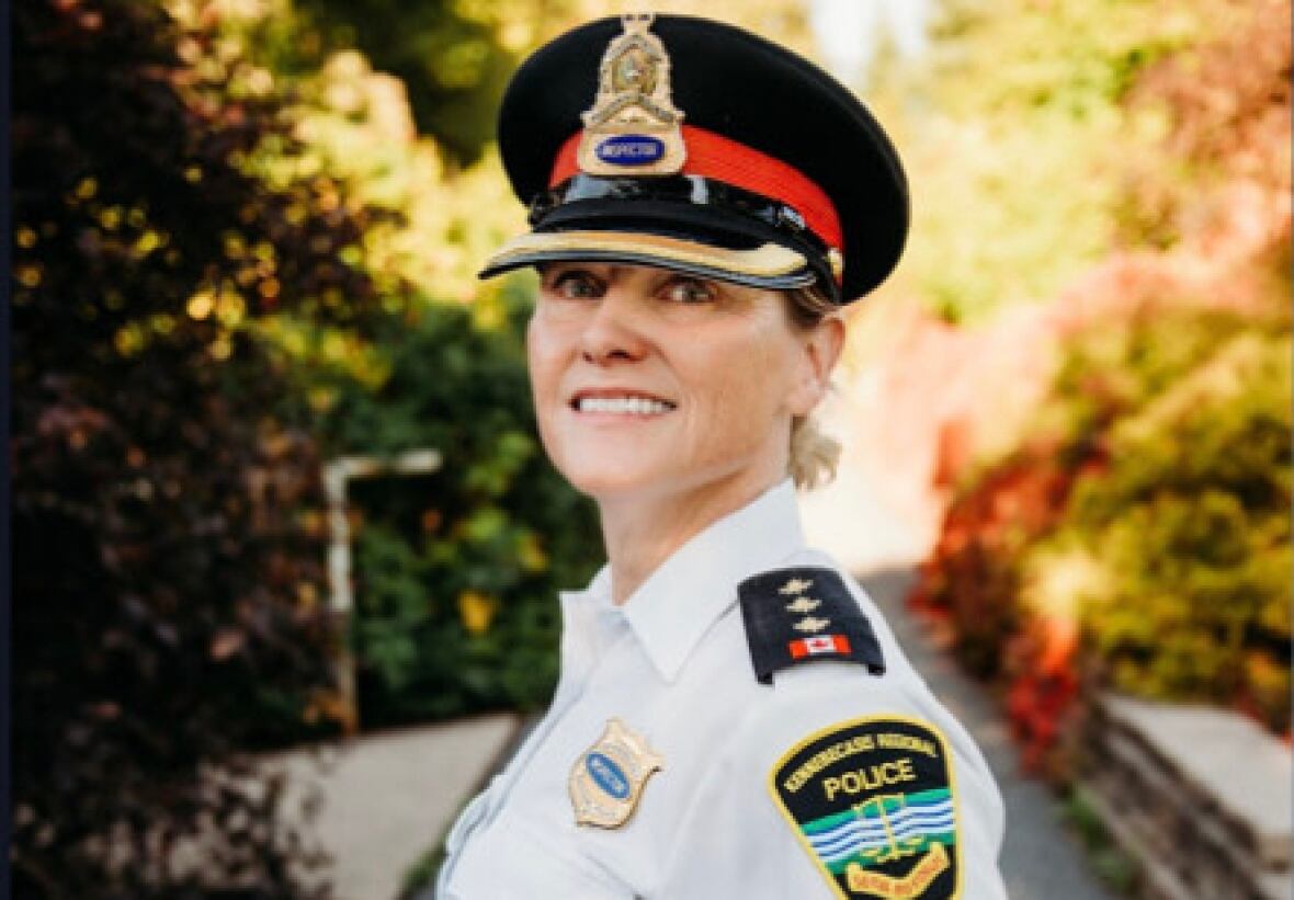 A smiling woman dressed in a police uniform stands outside on a road in front of trees.