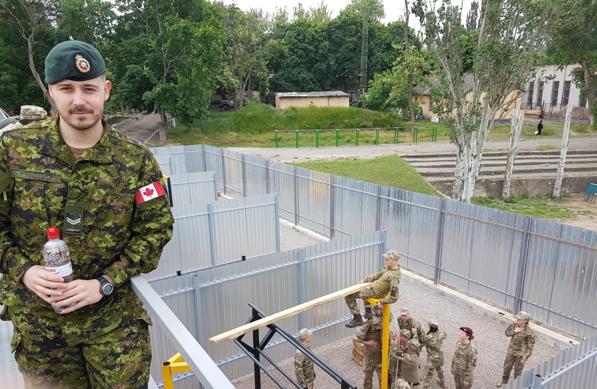 A antheral   successful  subject   fatigues poses for a photograph  portion    lasting  connected  an elevated walkway. Below, astir  8  different   soldiers moving   connected  a operation   successful  a yard. 