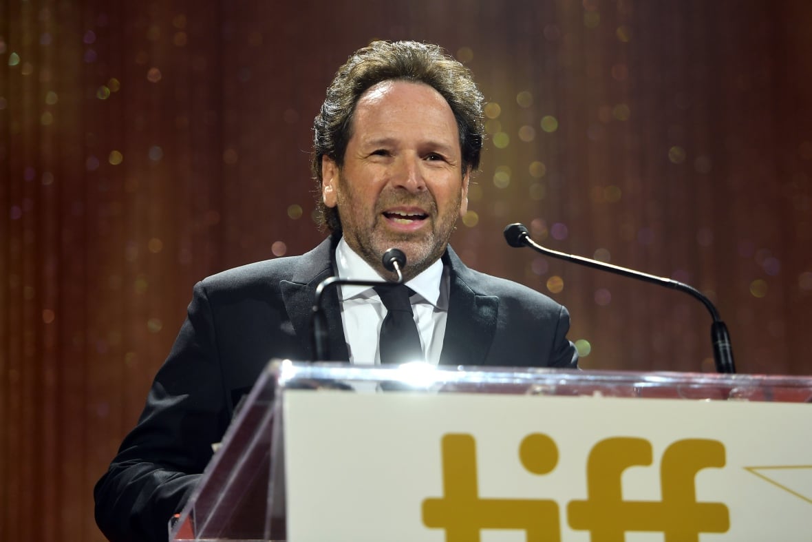 A man wearing a black suit stand after a podium with a word "Deception" before. A man smiles while speaking about microphones in podium and has brought a talk