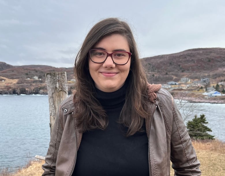 A woman wearing a black turtleneck and a brown jacket stands with a small harbour in the backdrop