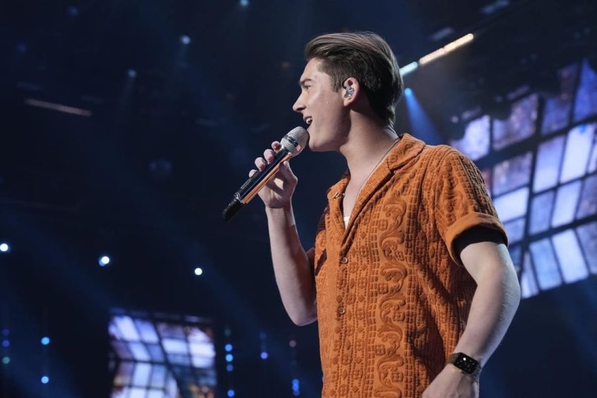 A man in an orange shirt sings on a stage lit up purple