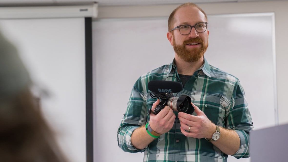 Bearded man with glasses stands in front of white board holding DSLR camera with microphone attachment.