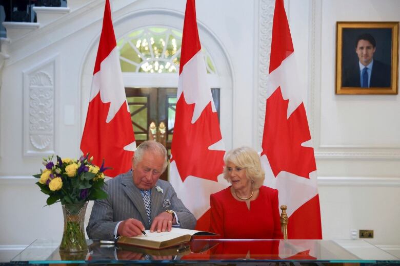 Two people sit at a table as one signs a guest book, with Canadian flags in the background.