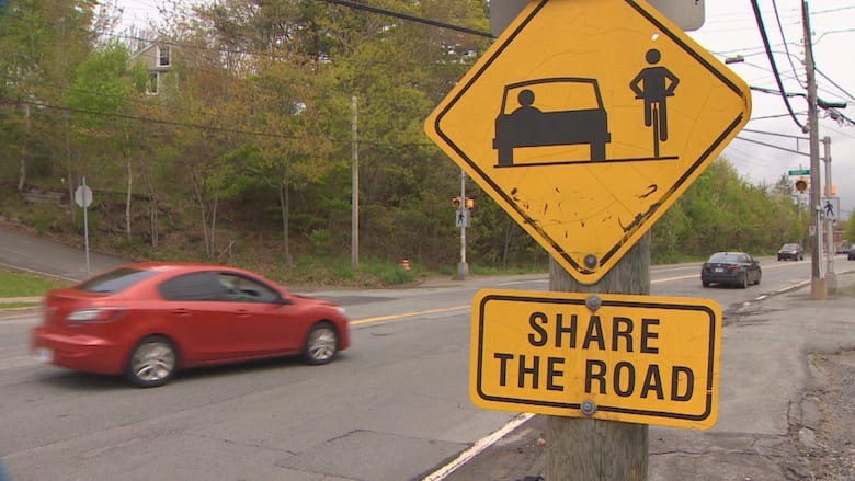 A road-side sign with a picture of a car and bike that reads "share the road." A car driving by is pictured in the background behind it.