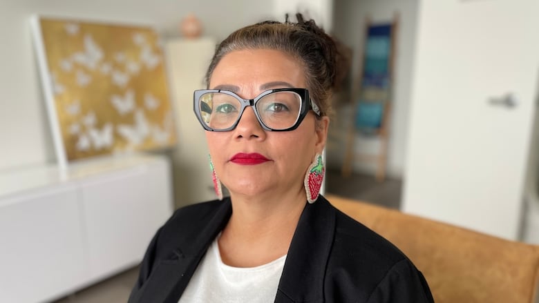 A woman in glasses, wearing strawberry-shaped beaded earrings, looks at the camera.