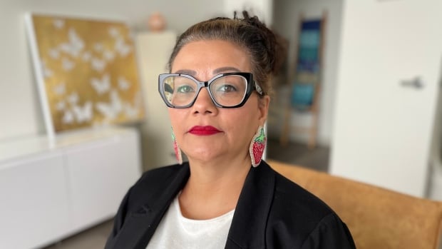 A woman in glasses, wearing strawberry-shaped beaded earrings, looks at the camera.