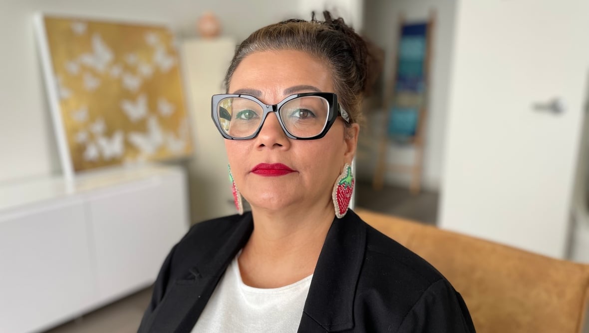 A woman in glasses, wearing strawberry-shaped beaded earrings, looks at the camera.