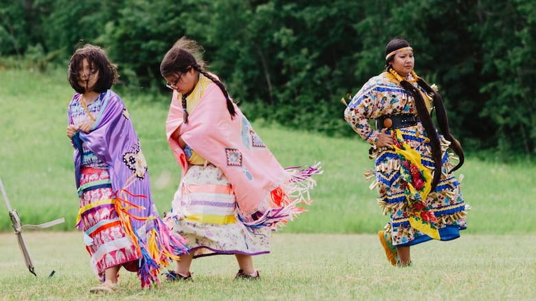 Three young women in their regalia dance during a powwow on a green field.