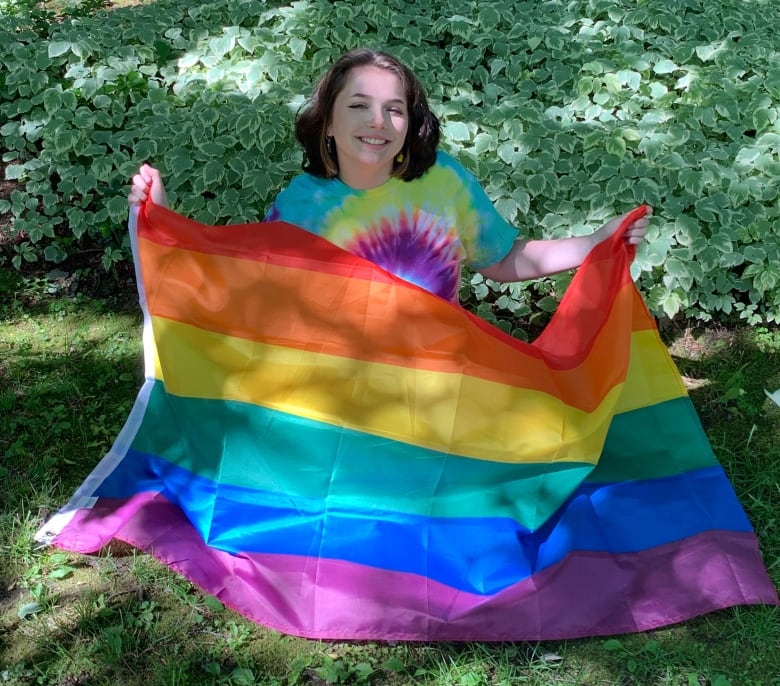 A student is waving a rainbow flag, sitting on the grass.